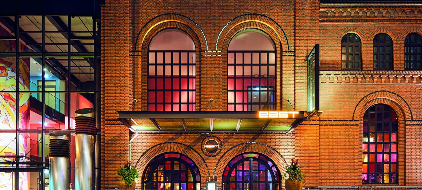 Historisches Gebäude aus rotem Backstein mit großen, bunt beleuchteten Bogenfenstern und Topfpflanzen am Eingang.