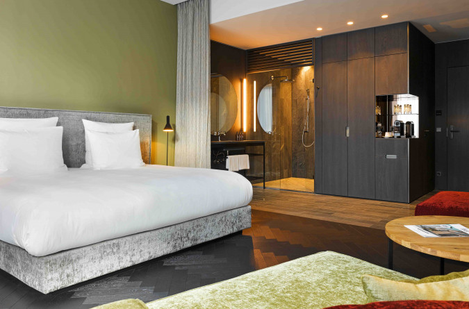 Modernes Zimmer in east Hotel mit einem bequemen Bett, weißer Bettwäsche und einer grünen Akzentwand. Ein offenes Badezimmer mit ebenerdiger Dusche und hinterleuchteten Spiegeln ist sichtbar. Zwei rote Stühle und ein runder Holztisch stehen im Vorderg
