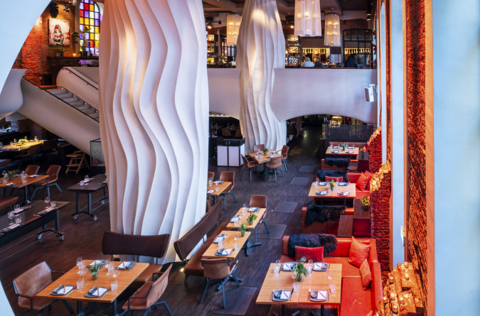 The modern restaurant interior of east with tall, wavy white columns, wooden dining tables, brown chairs, red seat cushions, large windows and warm ambient lighting.