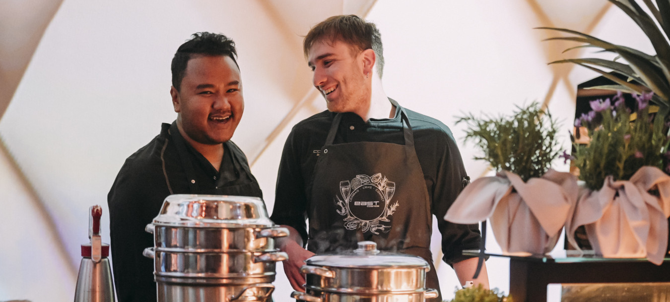Two chefs smile and chat behind a counter with stacked pots, herbs, and bowls at a modern kitchen setup.