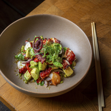 Eine Keramikschüssel mit einem bunten Salat aus Avocado, Oktopus, roten Zwiebeln und Gemüse steht auf einem Holztisch. Auf der rechten Seite der Schüssel liegen Essstäbchen.