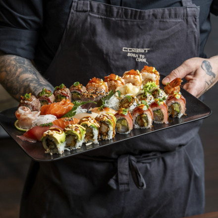 Eine Person hält ein Tablett mit verschiedenen Sushi-Rollen, belegt mit verschiedenen Zutaten wie Lachs, Avocado und Rogen. Die Person trägt eine dunkle Schürze und hat einen sichtbaren tätowierten Arm.