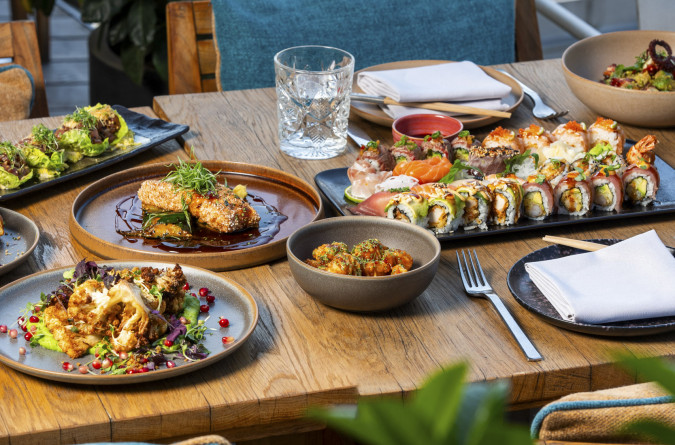 Ein Holztisch im coast by east Restaurant mit verschiedenen Gerichten, darunter Sushi-Rollen, gegrillter Fisch mit Gemüse, gebratenes Gemüse, ein kleiner Salat und ein Wasserglas neben Servietten. Im Hintergrund sind eine Pflanze und ein Teil eines Stuh