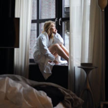 Junge blonde Frau im Bademantel sitzt auf Fensterbank und sieht verträumt aus dem Fenster