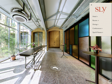 Ein heller, moderner Flur im sly Hotel mit großen Glasfenstern, bunten Glaspaneelen und hohen Türen. Topfpflanzen und Getränke stehen auf hohen Tischen. Eine Menüeinblendung zeigt die Navigation der Roomtour.