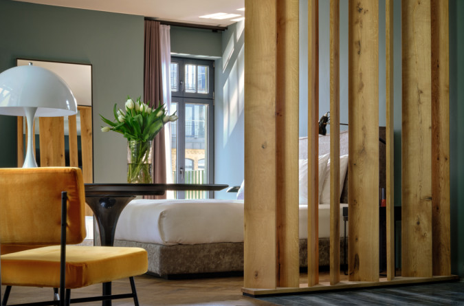 Gemütliches Hotelzimmer im Sly Hotel mit Trennwand. Es verfügt über ein bequemes Bett, eine Trennwand aus Holzlatten, einen Beistelltisch mit einer Vase mit weißen Blumen, einen gelben Stuhl und eine große Stehlampe. Sonnenlicht fällt durch ein gro