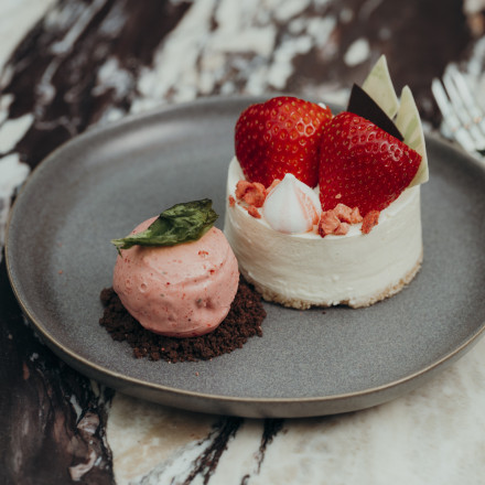 Ein Dessertteller mit einem runden Käsekuchen, garniert mit Erdbeerscheiben und Schokoladensplittern, daneben eine Kugel rosa Eis auf einem Streuselboden, garniert mit einem grünen Blatt. Der Teller steht auf einer Marmoroberfläche.