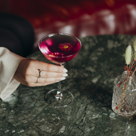 Eine Frauenhand mit weißem Nagellack und Schmuckringen hält ein Martini-Glas mit dunkelrotem Cocktail, garniert mit einer Blume. Das Getränk steht auf einem grünen Marmortisch neben einer kleinen Vase mit getrockneten Blumen.